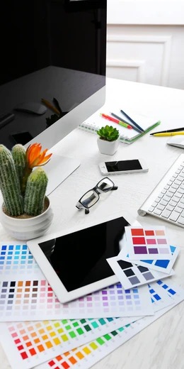 Mesa escritorio con un ordenado, paletas de colores, una tableta, móvil y unas plantas.